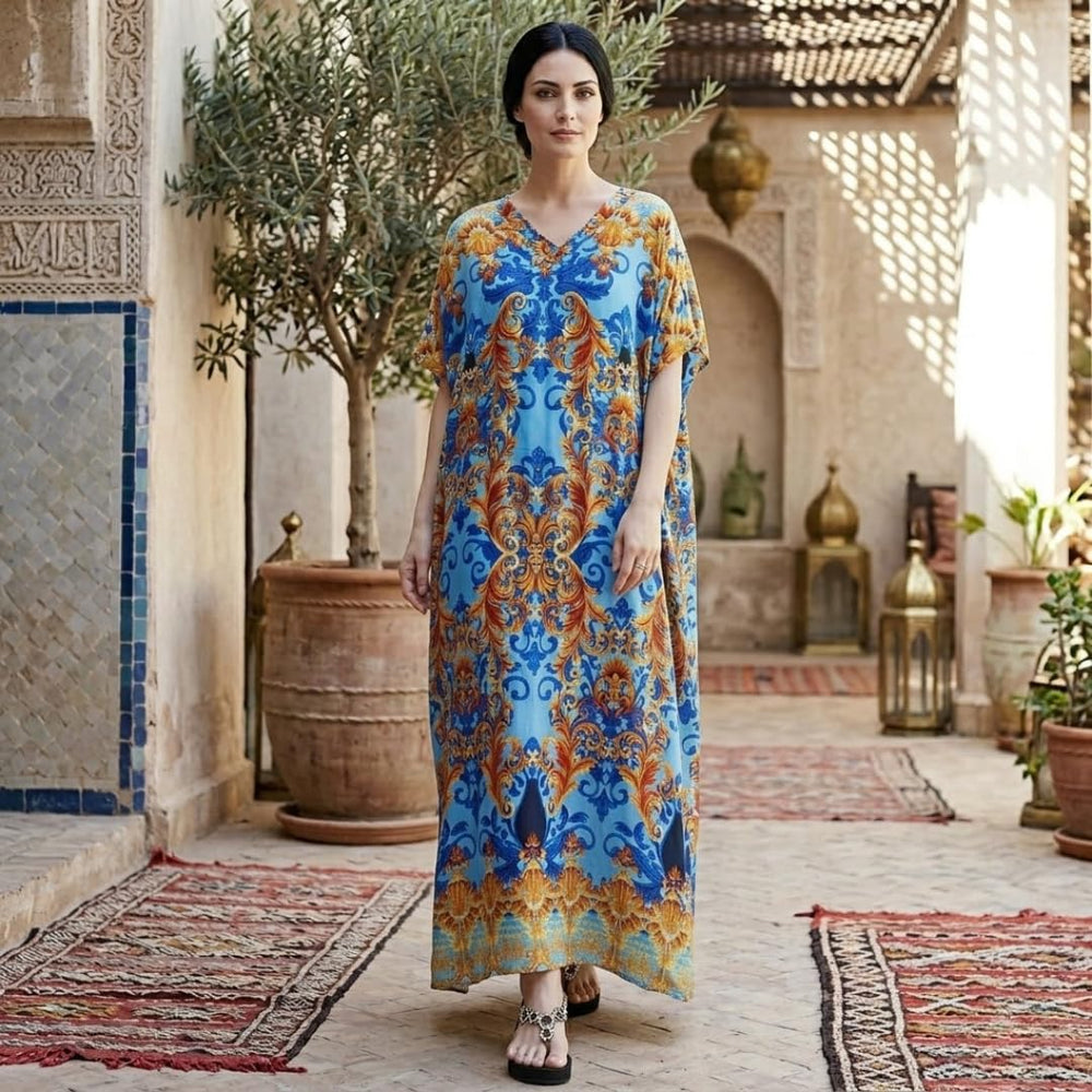 Model wearing a light blue floral print kaftan dress with intricate orange and gold baroque patterns, styled as a maxi-length resortwear cover-up in a Mediterranean setting.